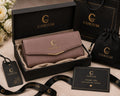 A Corium wallet presented in an open black gift box with branded packaging, including a black shopping bag, drawstring pouch, hangtag, card, and ribbon, arranged on a beige surface with white flowers at the side.